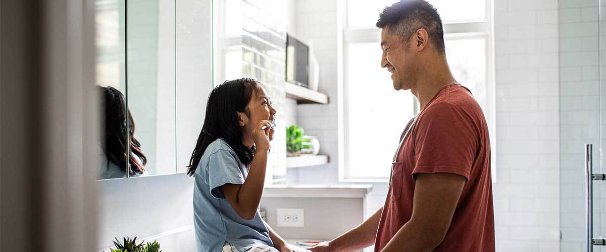 Little girl brushing teeth while dad looks at her and smiles